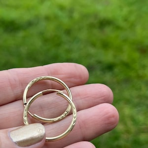 Puede incluir: Un par de pendientes de aro dorados con un diseño texturizado. Los pendientes se sostienen en una mano sobre un fondo verde borroso. Los aros tienen un cierre de bisagra.