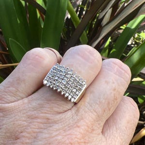 May include: A wide silver-toned ring featuring a multi-row design densely set with numerous small, clear, round-cut stones. The band has a flat, rectangular face that spans across the finger, showcasing a grid-like arrangement of sparkling stones.