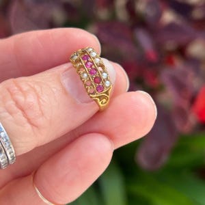 May include: A vintage gold ring featuring a row of vibrant red gemstones and small white pearls. The ring has an ornate design with delicate filigree details. The ring is held up close, showcasing its intricate craftsmanship and the sparkle of the gemstones.