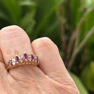 May include: A gold ring featuring a series of marquise-cut, deep red gemstones alternating with smaller, round, clear gemstones. The ring is set on a gold band and is shown on a person's finger.