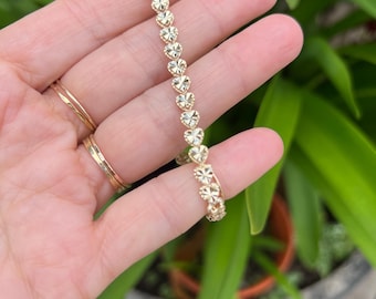May include: A gold bracelet composed of a continuous chain of small, faceted heart-shaped links. Each heart link features a textured, reflective surface that catches the light, creating a shimmering effect along the entire length of the jewelry piece.