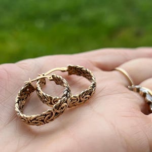 Może przedstawiać: Para kolczyków w kolorze złota z misternym, tkanym wzorem. Kolczyki są prezentowane na dłoni, z rozmytym zielonym tłem. Kolczyki mają klasyczny, elegancki styl.