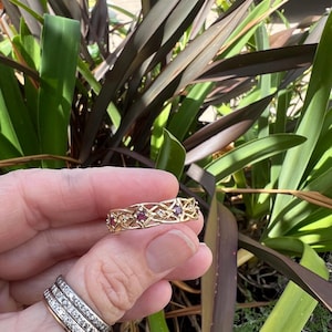 Może przedstawiać: Złoty pierścionek o skomplikowanym ażurowym wzorze, z małymi czerwonymi kamieniami szlachetnymi i przezroczystymi kamieniami akcentującymi. Pierścionek jest trzymany w dłoni, z innymi pierścionkami widocznymi na palcu. Tło zawiera zielone i ciemnoczerwone liście.