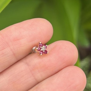 May include: A gold stud earring shaped like a five-petaled flower, featuring five round red rubies as petals and a small clear diamond at the center. The earring has a metal post backing and is held between two fingers against a blurred green background.