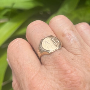 May include: A gold signet ring featuring an octagonal face with an engraved monogram of the letters J and C in a cursive script. The band displays intricate etched detailing along the sides, providing a textured finish to the metal surface.