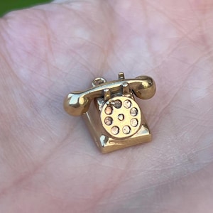 Pode incluir: Um pequeno pingente de telefone dourado. O pingente apresenta um design clássico de discagem rotativa com um fone e uma base. O pingente é provavelmente feito de metal e é mostrado em um fundo desfocado.