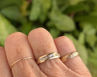 May include: Three gold rings worn on fingers. The first is a thin, plain gold band. The second is a wider gold band featuring a channel setting with five small, clear, round-cut stones. The third is a gold band with an engraved floral pattern.