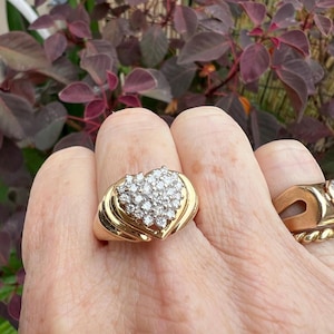 May include: A gold ring with a heart-shaped cluster of small, clear gemstones. The ring has a wide band and a detailed setting. The gemstones are arranged to form a heart shape. The ring is worn on a finger.