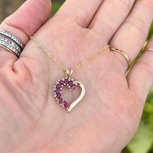 May include: A gold-colored heart-shaped pendant necklace. The pendant is adorned with a cluster of oval-cut, deep red gemstones and small white gemstones. The necklace is displayed on a hand.