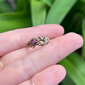 May include: A gold-toned metal ring featuring a textured, scale-like design that wraps around the finger. The ring is accented with a small, round, deep red gemstone set securely within the intricate, organic metalwork of the band.