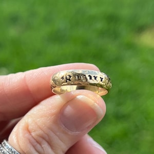 Może przedstawiać: Złoty pierścionek z grawerowanym wzorem. Pierścionek ma zaokrąglony kształt i wydaje się być wykonany z błyszczącego, żółtego metalu. Pierścionek jest trzymany przez palce osoby na rozmytym zielonym tle.