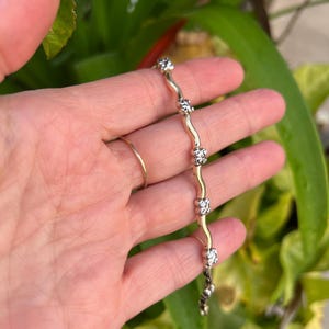 May include: A gold and silver bracelet with a wavy design. The bracelet features a series of silver-colored, flower-shaped accents connected by a gold-colored, serpentine chain. The bracelet is held in a hand, with a gold ring on a finger.