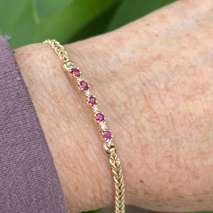 May include: A gold bracelet with a woven chain design. The bracelet features a row of small, round, red gemstones and clear gemstones. The bracelet is worn on a wrist with a purple sleeve.
