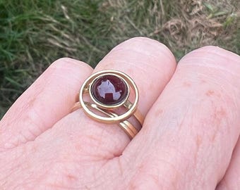 May include: A gold-colored ring with a dark red gemstone set in a circular design. The ring has a double band and a unique, openwork setting around the stone. The gemstone is a deep, rich color, contrasting with the gold metal.