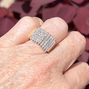 May include: A rectangular ring with a gold band and a surface covered in small, sparkling stones. The ring is worn on a finger, with a gold band visible on the adjacent finger. The background is a blurred mix of red and pink.