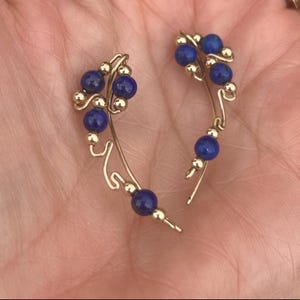 May include: Ear climbers with a gold-tone wire design. Each earring features multiple deep blue, round beads and small gold-tone accents. The earrings curve upwards, designed to be worn along the ear.