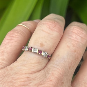 Puede incluir: Un anillo de banda de oro con piedras preciosas rojas alternas y pequeñas piedras claras. El anillo se muestra sobre un fondo verde borroso, con un dedo y parte de una mano visibles. El anillo es una joya.