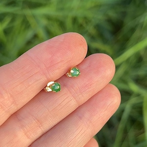 May include: A pair of small stud earrings featuring round, polished green jade stones set in gold-toned metal prongs. Each earring has a simple, circular design with a smooth, cabochon-cut green gemstone securely held by a metallic base.