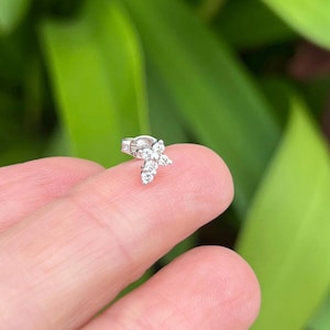May include: A small, silver-colored stud earring with a floral design. The earring features multiple clear, sparkling stones arranged in a cluster, creating a delicate, eye-catching piece of jewelry. The earring is shown against a blurred green background.