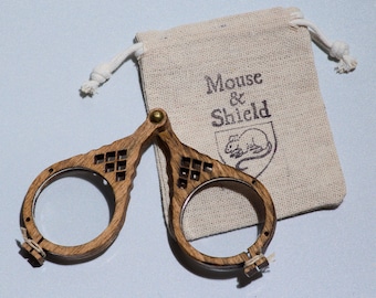 May include: A pair of folding wooden spectacles with circular frames and a brass hinge, accompanied by a small beige drawstring pouch featuring the printed text Mouse & Shield above a line drawing of a mouse inside a shield emblem.