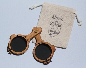 May include: A pair of folding wooden spectacles with dark circular lenses and a brass hinge, accompanied by a beige drawstring pouch featuring the printed text Mouse & Shield above a graphic illustration of a mouse inside a shield emblem.