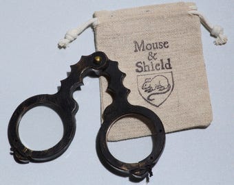 May include: A dark brown, hinged wooden tool with two circular openings and a brass-colored pivot pin, accompanied by a beige drawstring pouch featuring the printed text Mouse & Shield above a line drawing of a mouse inside a shield emblem.