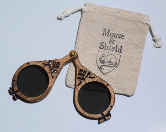 May include: A pair of folding spectacles with dark circular lenses and a brown wooden frame featuring intricate cut-out details, connected by a brass hinge. Beside the glasses is a small beige drawstring pouch with the text Mouse & Shield.
