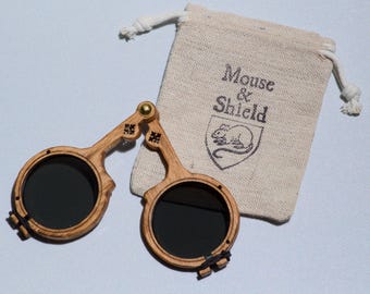 May include: A pair of folding spectacles with circular wooden frames and dark lenses, featuring a brass hinge at the bridge. Beside the glasses is a beige drawstring pouch made of fabric, printed with the text Mouse & Shield and a mouse icon.