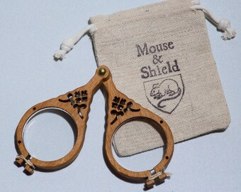 May include: A pair of folding wooden magnifying glasses with circular frames and decorative cutouts, joined by a brass rivet. Beside the glasses is a small, beige drawstring pouch made of woven fabric, featuring the text Mouse & Shield.