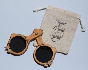 May include: A pair of folding wooden spectacles with dark circular lenses and a brass hinge, accompanied by a small beige drawstring pouch featuring the printed text Mouse & Shield above a graphic illustration of a mouse inside a shield emblem.