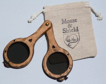 May include: A pair of folding wooden spectacles with dark circular lenses and a brass hinge, accompanied by a small beige drawstring pouch featuring the printed text Mouse & Shield above a graphic illustration of a mouse inside a shield emblem.