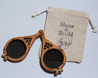 May include: A pair of folding wooden spectacles with dark circular lenses and a geometric cutout design on the frame, accompanied by a small beige drawstring pouch featuring the printed text Mouse & Shield and a graphic of a mouse inside a shield.