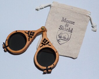May include: A pair of folding wooden sunglasses with dark circular lenses and intricate cutout floral patterns on the frames, connected by a brass hinge. Beside the glasses is a small beige drawstring pouch with the text Mouse & Shield.