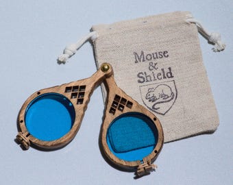 May include: A pair of hinged wooden spectacles with circular blue lenses and a decorative geometric cutout pattern on the frame. Beside the spectacles is a beige drawstring pouch featuring the printed text Mouse & Shield and a mouse illustration.