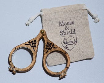 May include: A pair of folding wooden spectacles with circular frames and intricate cut-out floral details, joined by a brass rivet. Beside the glasses is a small beige fabric drawstring pouch featuring the text Mouse & Shield and a mouse icon.