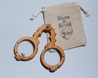 May include: A pair of folding wooden reading glasses with a +2.00 lens power sticker on the left lens. The glasses feature a serrated handle design and are accompanied by a beige drawstring fabric pouch printed with the text Mouse & Shield.