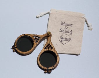 May include: A pair of folding wooden spectacles with dark circular lenses and a brass hinge, accompanied by a small beige drawstring pouch featuring the printed text Mouse & Shield above a crest containing an illustration of a mouse.
