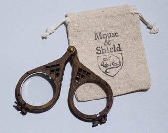 May include: A pair of dark brown wooden folding spectacles with circular frames and a brass hinge, accompanied by a small beige canvas drawstring pouch featuring the printed text Mouse & Shield and a graphic illustration of a mouse inside a shield.
