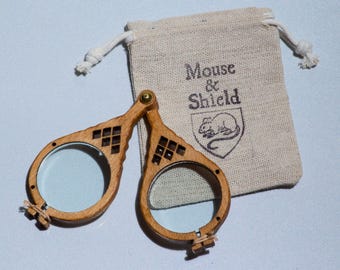 May include: A pair of folding spectacles with light brown wooden frames and circular lenses. The frames feature a decorative cutout pattern near the bridge. A small beige drawstring pouch is included, printed with the text Mouse & Shield.
