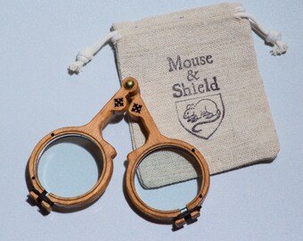 May include: A pair of folding spectacles with light brown wooden frames and clear lenses, connected by a brass hinge. The glasses are accompanied by a small, beige fabric drawstring pouch featuring the text Mouse & Shield and a mouse illustration.