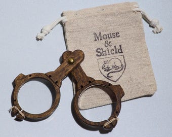 May include: A pair of dark brown wooden spectacles with circular frames and a brass rivet hinge, accompanied by a beige canvas drawstring pouch featuring the printed text Mouse & Shield above a line drawing of a mouse inside a shield emblem.
