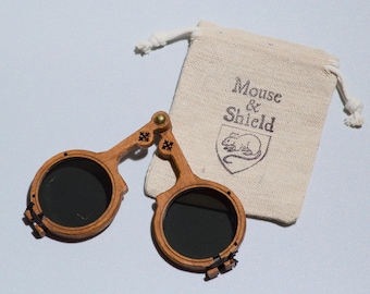May include: A pair of folding wooden spectacles with dark circular lenses and a brass hinge, accompanied by a small beige drawstring pouch featuring the printed text Mouse & Shield above a graphic illustration of a mouse inside a shield emblem.