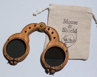 May include: A pair of wooden spectacles with dark circular lenses and a hinged bridge, accompanied by a beige fabric drawstring pouch featuring the printed text Mouse & Shield above a line drawing of a mouse inside a shield emblem.