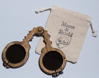 May include: A pair of folding spectacles with dark circular lenses and a brown wooden frame, featuring a brass hinge at the bridge. Beside the glasses is a small beige drawstring pouch with the text Mouse & Shield and a mouse illustration.