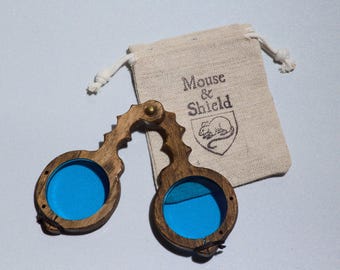 May include: A pair of folding wooden spectacles with circular frames and bright blue lenses, featuring a dark brown wood grain finish and a brass hinge. Beside the spectacles is a beige drawstring pouch with the text Mouse & Shield printed.