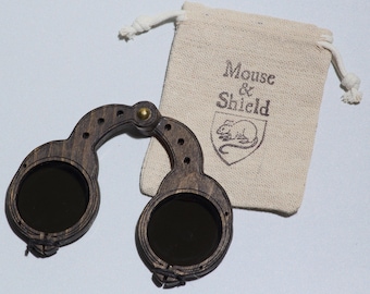 May include: A pair of dark brown wooden folding spectacles with circular dark lenses and a brass hinge, accompanied by a small beige drawstring pouch featuring the printed text Mouse & Shield and a graphic illustration of a mouse inside a shield.