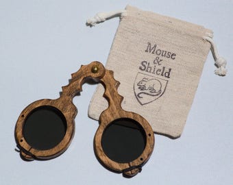 May include: A pair of folding spectacles with a dark brown wood frame and circular black lenses, featuring a brass hinge at the bridge. Beside the glasses is a small beige drawstring pouch with the text Mouse & Shield and a mouse illustration.
