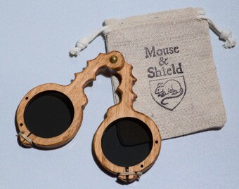 May include: A pair of folding wooden spectacles with dark circular lenses and a textured, carved frame. The glasses are accompanied by a small, beige drawstring pouch made of woven fabric, featuring the printed text Mouse & Shield with a mouse icon.