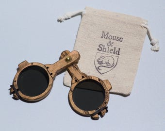 May include: A pair of folding wooden spectacles with dark circular lenses and a brass hinge, accompanied by a small beige drawstring pouch featuring the printed text Mouse & Shield and a graphic illustration of a mouse inside a shield emblem.