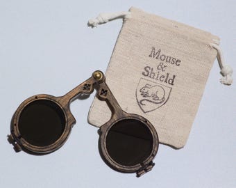May include: A pair of dark-tinted, circular wooden spectacles with a hinged bridge and a small brass screw, accompanied by a beige drawstring pouch featuring the printed text Mouse & Shield above a graphic of a mouse inside a shield emblem.